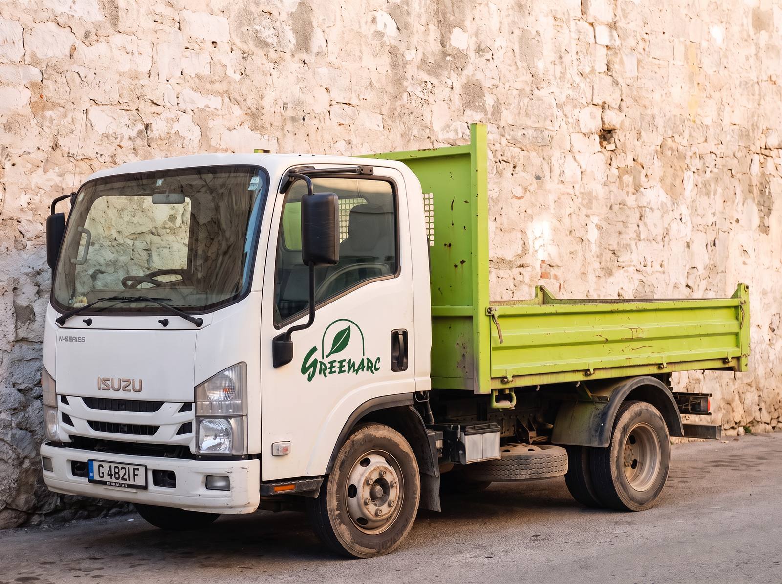 White Isuzu tipper truck with a lime-green tipper body and the Greenarc logo on the door, parked against a limestone wall.