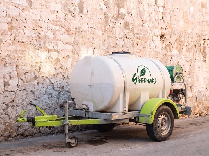 White 1,000 litre water bowser on a trailer with lime-green mudguards and the Greenarc logo on the tank, against a limestone wall.